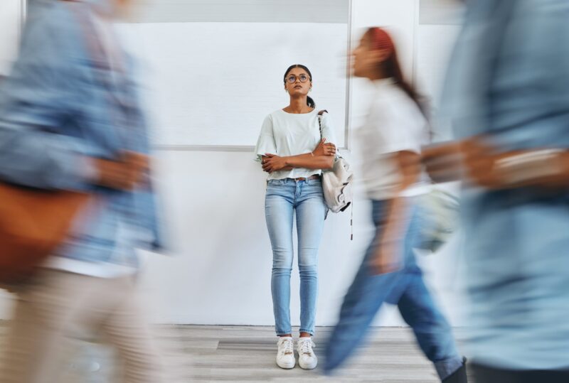 A young woman stands near a wall, looking upward, while people cross in front of her